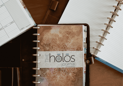 A weekly planner layout shown open on a wooden desk, alongside a closed brown leather folio containing a spiral-bound journal titled 'the holos journal'.